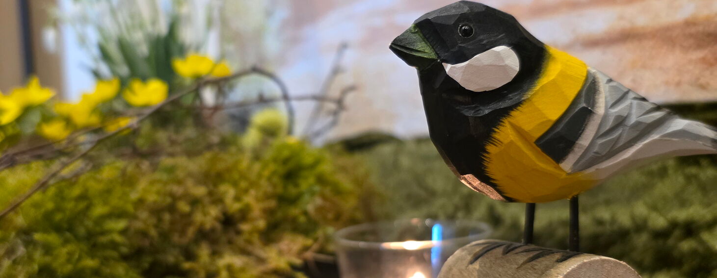 Ausschnitt einer Dekoration für eine Trauerfeier Eine Kohlmeise aus Holz sitzt auf einer Rinde. Im Hintergrund brennt ein Teelicht und ein Mooskranz mit Frühblühern ist zu sehen,